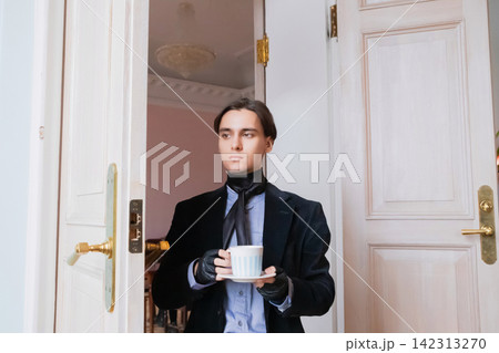 Aristocrat man standing in doorway holding cup of tea. Elegant gentleman in black jacket and leather glove lifestyle portrait. Classic luxury interior background. Rich style concept. Aristocrat man standing in doorway holding cup of tea. Elegant gentleman in black jacket and leather glove lifestyle portrait. Classic luxury interior background. Rich style concept. 142313270