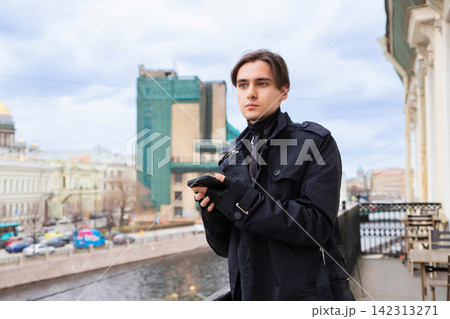Young man standing on a city balcony holding a smartphone. Stylish guy in a black trench coat looking away while using a mobile phone. Urban lifestyle and communication in a city environment. 142313271