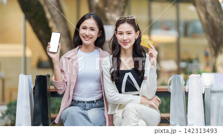 Asian women holding smartphone and credit card while sitting with shopping bags outdoors 142313493