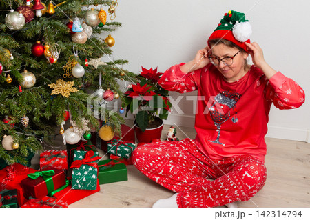 A 52-year-old woman sits on the floor next to a Christmas tree. She wears festive clothes and holds a decoration as she looks at gifts on December 25th. The room is filled with holiday cheer A 52-year-old woman sits on the floor next to a Christmas tree. She wears festive clothes and holds a decoration as she looks at gifts on December 25th. The room is filled with holiday cheer 142314794