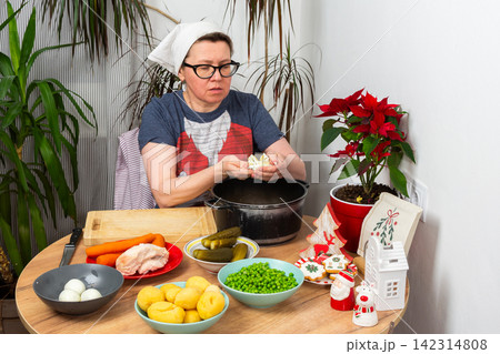 A woman prepares Olivier salad, Russian salad, in her kitchen, slicing boiled eggs and arranging vegetables on the table. This traditional dish is made for Christmas and New Year gatherings A woman prepares Olivier salad, Russian salad, in her kitchen, slicing boiled eggs and arranging vegetables on the table. This traditional dish is made for Christmas and New Year gatherings 142314808