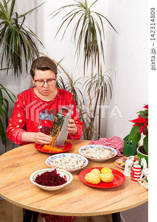 A woman grates carrots in the kitchen for making herring under fur coat salad, the traditional Ukrainian or Russian Christmas salad Shuba. Nearby are bowls with beets, potatoes, and a festive plant A woman grates carrots in the kitchen for making herring under fur coat salad, the traditional Ukrainian or Russian Christmas salad Shuba. Nearby are bowls with beets, potatoes, and a festive plant 142314809