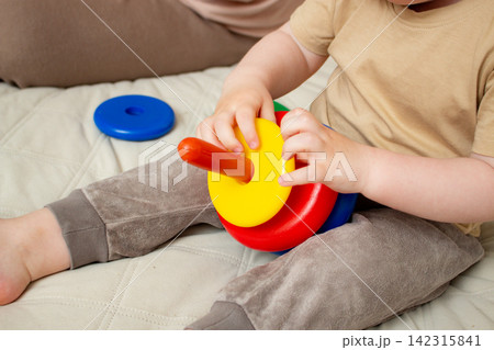 Mother helping child build pyramid on cozy bed. Educational play with shapes and colors. Parent interaction into child game. Family bonding concept Mother helping child build pyramid on cozy bed. Educational play with shapes and colors. Parent interaction into child game. Family bonding concept 142315841