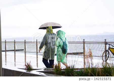 Travelers wearing raincoats with umbrellas walking through wet streets exploring new places under rain and gray sky. Bad weather not ruin adventure concept Travelers wearing raincoats with umbrellas walking through wet streets exploring new places under rain and gray sky. Bad weather not ruin adventure concept 142315871