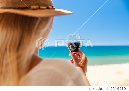 Woman in straw hat holding glass of red wine while enjoying turquoise sea view and relaxing on sunny beach vacation Woman in straw hat holding glass of red wine while enjoying turquoise sea view and relaxing on sunny beach vacation 142316910