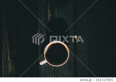 Top view of a cup of black coffee on a dark wooden table, with strong shadows and minimalist composition. The moody still life evokes quiet mornings, solitude, simplicity and slow living. Top view of a cup of black coffee on a dark wooden table, with strong shadows and minimalist composition. The moody still life evokes quiet mornings, solitude, simplicity and slow living. 142317091