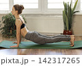 Woman performs yoga on a green mat in a sunny room with plants during the day 142317265