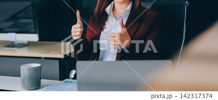 Smiling asian businesswoman talking on phone and using laptop while working at office desk 142317374