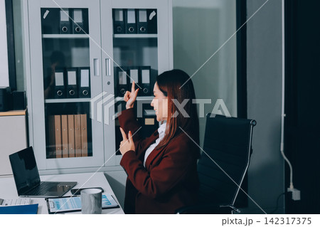 Smiling asian businesswoman talking on phone and using laptop while working at office desk 142317375