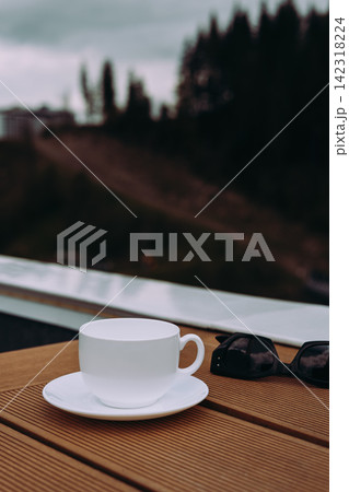 White cup and saucer on a wooden terrace beside sunglasses, with a blurred green hillside in the background. Peaceful outdoor scene evokes relaxation, travel, leisure and slow living in nature. today. White cup and saucer on a wooden terrace beside sunglasses, with a blurred green hillside in the background. Peaceful outdoor scene evokes relaxation, travel, leisure and slow living in nature. today. 142318224