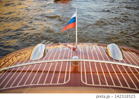 the glossy stern of a vintage pleasure boat at sunset, the boat in motion, bubbling water from under the boat, the flag of Russia fluttering in the wind, Neva river, varnished wood, chrome details the glossy stern of a vintage pleasure boat at sunset, the boat in motion, bubbling water from under the boat, the flag of Russia fluttering in the wind, Neva river, varnished wood, chrome details 142318525