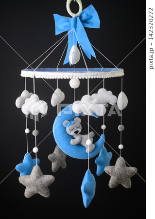 A handmade blue and white nursery cot mobile with a sleeping teddy bear resting on a crescent moon, surrounded by soft clouds, stars, and hanging ornaments against a dark backdrop 142320272