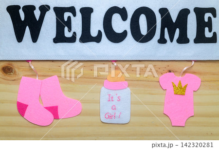 A handmade felt welcome sign decorated with pink baby clothing, tiny socks, a baby bottle, and a crown motif on a wooden background. Celebrates the arrival of a baby girl A handmade felt welcome sign decorated with pink baby clothing, tiny socks, a baby bottle, and a crown motif on a wooden background. Celebrates the arrival of a baby girl 142320281