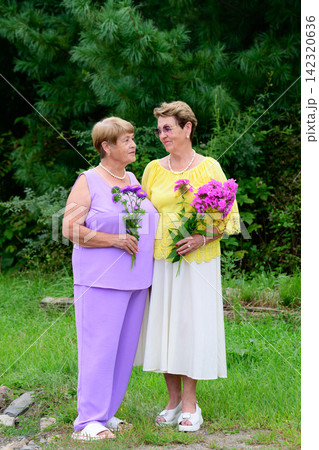 Full length portrait of two senior russian sisters looking at each other and smiling with flower bouquets standing in a garden in Russia 142320636