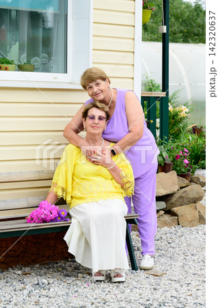 Authentic portrait of two senior russian sisters 80 and 75 years old sitting on a bench near a house porch in a summer garden in Russia Authentic portrait of two senior russian sisters 80 and 75 years old sitting on a bench near a house porch in a summer garden in Russia 142320637