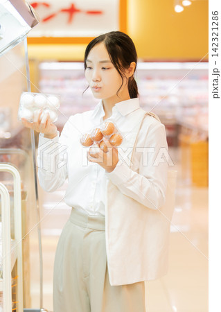 スーパーの卵売り場で白い卵と茶色い卵を比較して選ぶ若い女性 スーパーの卵売り場で白い卵と茶色い卵を比較して選ぶ若い女性 142321286