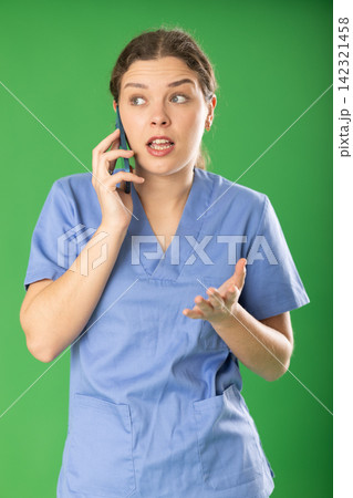 Professional female doctor wearing blue medical clothing convincingly and emotionally talking to patient by smartphone standing against green screen 142321458