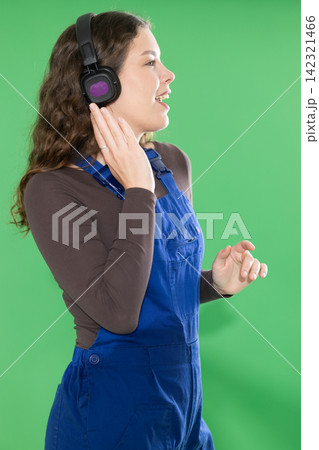 Female worker in a blue overalls happily listens to music on headphones and dances against green background Female worker in a blue overalls happily listens to music on headphones and dances against green background 142321466