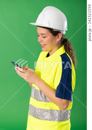 Polite girl worker wearing high visibility polo and hard hat scrolling smartphone standing against green screen Polite girl worker wearing high visibility polo and hard hat scrolling smartphone standing against green screen 142321559