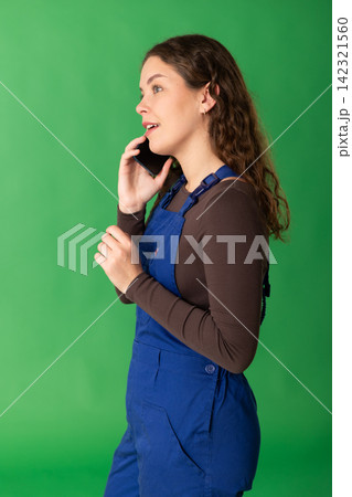 Portrait of girl worker dressed in blue work overalls talking to client by smartphone standing against green screen Portrait of girl worker dressed in blue work overalls talking to client by smartphone standing against green screen 142321560