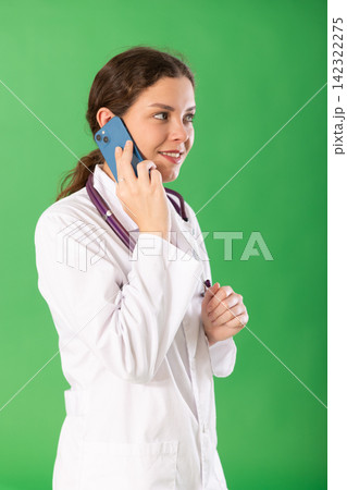 Young female doctor in white coat with stethoscope talking on mobile phone on green background Young female doctor in white coat with stethoscope talking on mobile phone on green background 142322275