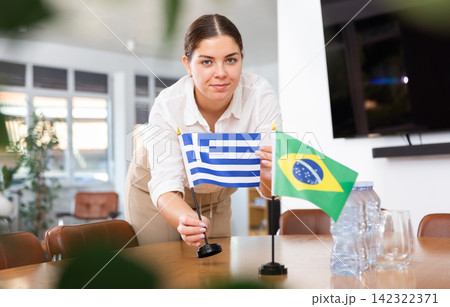 Positive young woman putting flag of Greece on the table with flag of Brazil in conference room 142322371
