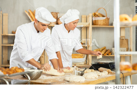 Two young bakers prepare dough in kitchen of bakery - together they knead dough, cut it with knife and weigh it on scales Two young bakers prepare dough in kitchen of bakery - together they knead dough, cut it with knife and weigh it on scales 142322477