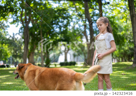 Young mixed race girl watching her dog exploring a sunny grass lawn surrounded by trees in the park. Young mixed race girl watching her dog exploring a sunny grass lawn surrounded by trees in the park. 142323416