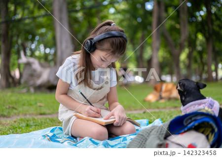 Young girl holding pencil writing or drawing in a notebook while a dog sits beside her in the park. Young girl holding pencil writing or drawing in a notebook while a dog sits beside her in the park. 142323458