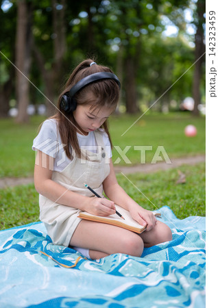 Young girl wearing headphones writing drawing in a notebook while sitting on picnic blanket in park Young girl wearing headphones writing drawing in a notebook while sitting on picnic blanket in park 142323459