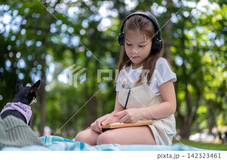 Young mixed race girl wearing headphones concentrates on writing drawing in notebook in the park. 142323461