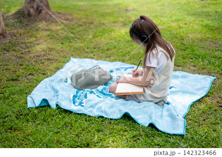 Side view of a young girl wearing headphones writes in a notebook while sitting on a blanket in park 142323466