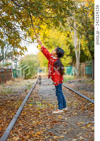 A happy young girl plays with a long wooden stick while standing under autumn trees near a railway track. 142324156