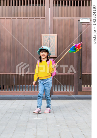 An adorable elementary school girl carrying a pink backpack stands by a large wooden gate holding a colorful pinwheel on a sunny morning. An adorable elementary school girl carrying a pink backpack stands by a large wooden gate holding a colorful pinwheel on a sunny morning. 142324197