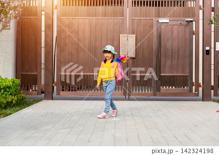 An adorable elementary school girl carrying a pink backpack stands by a large wooden gate holding a colorful pinwheel on a sunny morning. An adorable elementary school girl carrying a pink backpack stands by a large wooden gate holding a colorful pinwheel on a sunny morning. 142324198