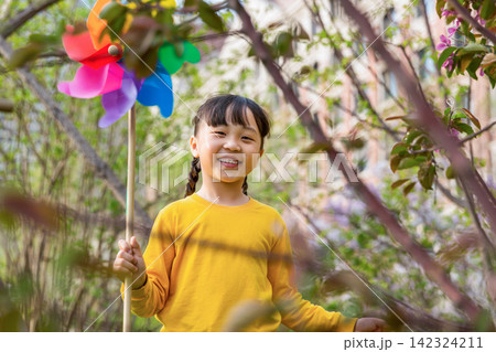 A joyful young girl in a yellow sweater looks back and smiles at the camera while holding a rainbow pinwheel in the park. A joyful young girl in a yellow sweater looks back and smiles at the camera while holding a rainbow pinwheel in the park. 142324211