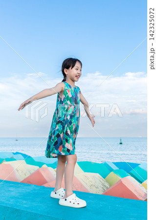 Happy little girl wearing a colorful dress standing on a blue seawall with open arms. Happy little girl wearing a colorful dress standing on a blue seawall with open arms. 142325072