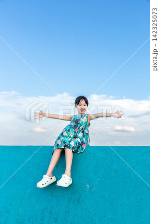 Smiling young girl sitting on a bright blue breakwater wall showing thumbs up. 142325073