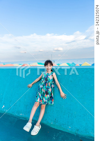Cheerful little girl leaning against a vibrant blue wall by the seaside. Cheerful little girl leaning against a vibrant blue wall by the seaside. 142325074
