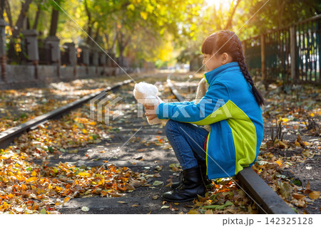 Side profile of a child sitting on a train rail playing with a white teddy bear on a sunny autumn day. 142325128