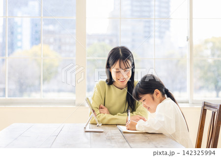 明るいリビングでタブレットを見ながらノートに向かう娘を優しく見守る母親 明るいリビングでタブレットを見ながらノートに向かう娘を優しく見守る母親 142325994