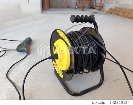 a black and yellow extension cord reel mounted on a black metal bracket against a rough concrete wall. Electrical tools a black and yellow extension cord reel mounted on a black metal bracket against a rough concrete wall. Electrical tools 142330124