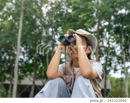 Cute Asian girl in safari hat looking through binoculars outdoors in forest 142332092