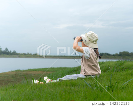 Little girl sitting on green grass by lake looking through binoculars Little girl sitting on green grass by lake looking through binoculars 142332133