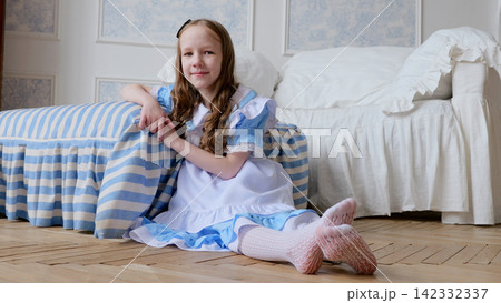Caucasian girl in costume seated on floor smiling beside bedspread, joyful party vibe, pastel palette with ribbons and lace, playful guest role, candid family celebration atmosphere 142332337