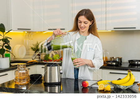 Middle-aged woman pouring fresh green smoothie from blender in home kitchen for detox breakfast diet Middle-aged woman pouring fresh green smoothie from blender in home kitchen for detox breakfast diet 142333056