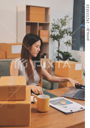 Startup SME small business entrepreneur of freelance Asian woman wearing apron using laptop and box to receive and review orders online to prepare to pack sell to customers, online sme business ideas. Startup SME small business entrepreneur of freelance Asian woman wearing apron using laptop and box to receive and review orders online to prepare to pack sell to customers, online sme business ideas. 142333810
