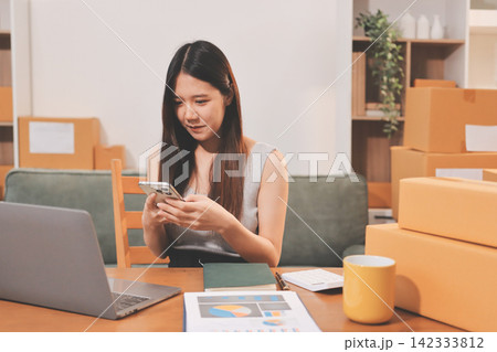 Startup SME small business entrepreneur of freelance Asian woman wearing apron using laptop and box to receive and review orders online to prepare to pack sell to customers, online sme business ideas. Startup SME small business entrepreneur of freelance Asian woman wearing apron using laptop and box to receive and review orders online to prepare to pack sell to customers, online sme business ideas. 142333812