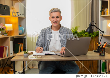 Middle-aged man on home table checking online bank transactions and writing expenses in a notebook 142334174