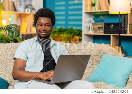 African American young man on home sofa typing on laptop with focused expression during remote work African American young man on home sofa typing on laptop with focused expression during remote work 142334261